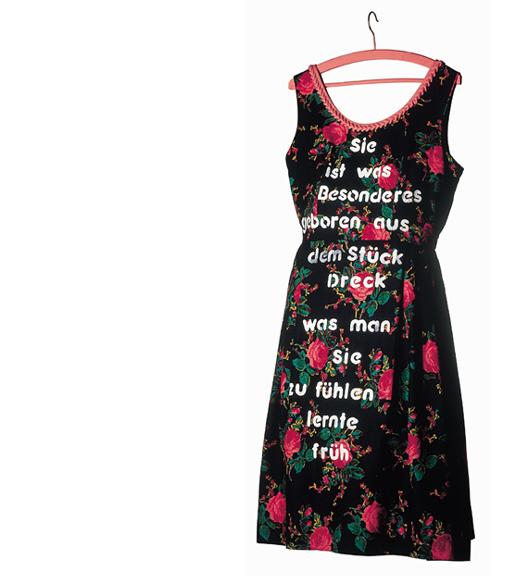 Ein Rosenkleid hängt an einem rosafarbenen Bügel. Das Kleid ist mit einem Text in großen weißen Buchstaben bedruckt: 